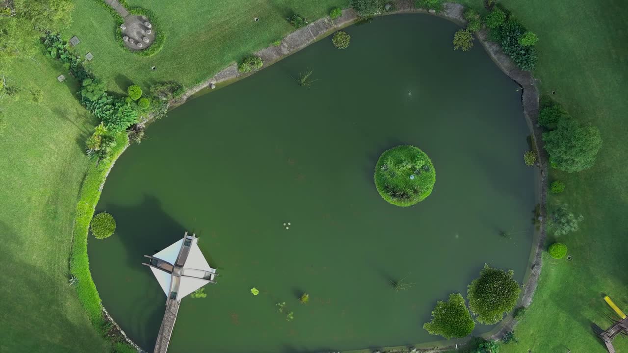 Aerial top-down view zooming out from an artificial lagoon with fish. Features a small green island in the center surrounded by a walking bridge and greenery. Guatemala.