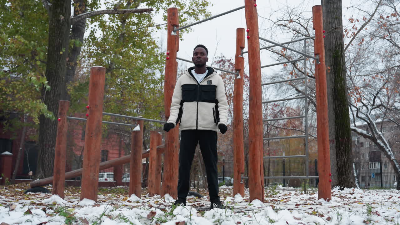 joven con guantes negros, chaqueta de invierno y pantalones negros realizando torsión de muñeca al aire libre, entorno de parque nevado con equipos de entrenamiento, hojas caídas y edificio residencial lejano