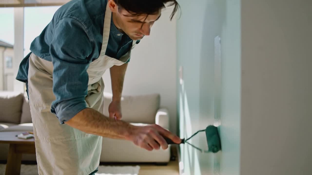 Placing roller, man wearing denim shirt, canvas apron, rolling paint down living room wall for DIY