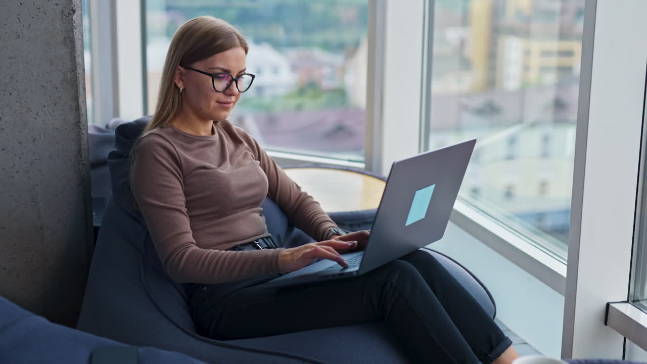 Good-looking blonde lady working on laptop sitting in bean bag chair. Lady distracts from work looking into big panoramic window she's sitting by.