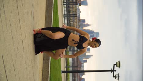 belly-dancer-outdoor-with-red-flower-in-her-fixed-hair-performing-moving-like-a-snake-waves-and-sharp-movements-black-dress-revealing-body-red-heels-great-city-London-in-the-background-fence-river-sky