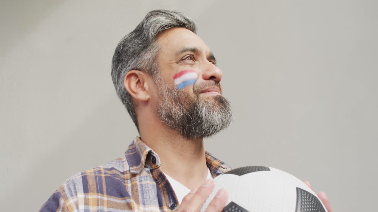 hombre biracial feliz con bandera de los países bajos en la mejilla sosteniendo un fútbol