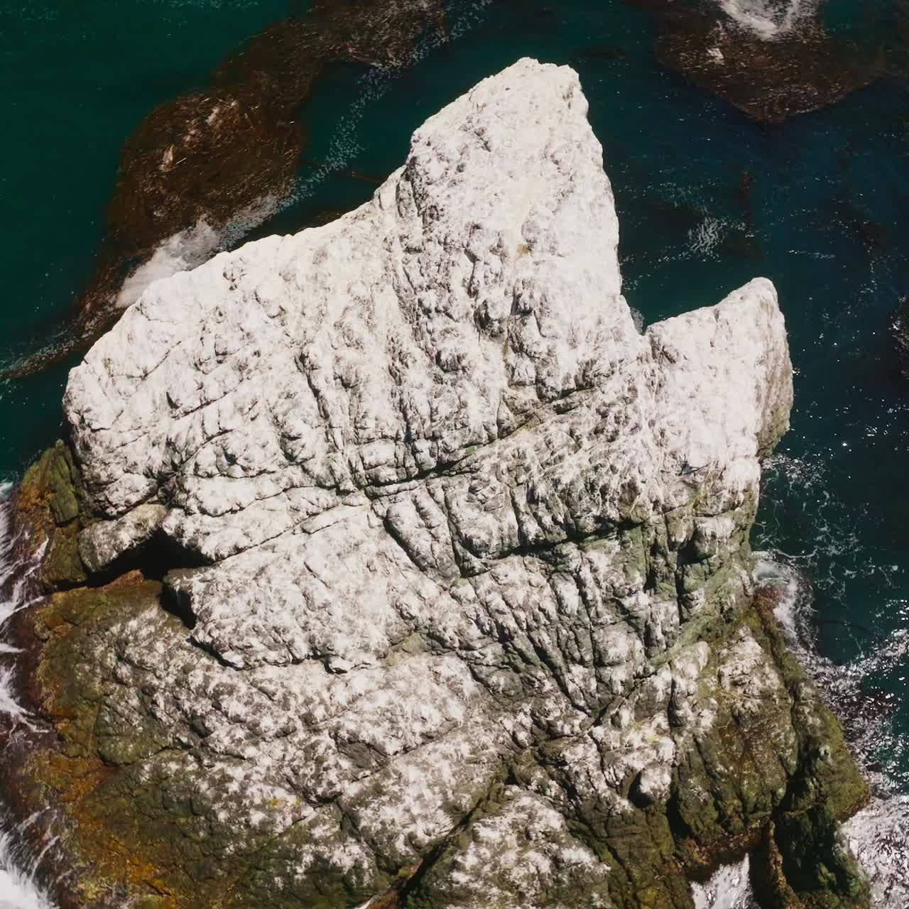 White rock peeping out from blue water of ocean on sunny day. Waves hitting the mount. Seaweeds floating on the surface of water. Top view