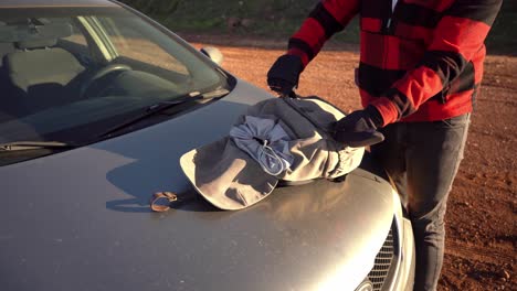 hombre desempacando un dron y un controlador de una mochila marrón en el capó de un auto, israel