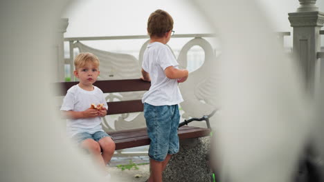 a child sits holding a sausage roll, looking thoughtful, while the older sibling stands next to the bench, touching his stomach, with both children in casual white t-shirts and denim shorts
