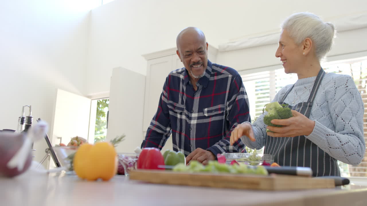 Casal sênior diverso feliz preparando vegetais, usando tablet na cozinha, com espaço para texto, em câmera lenta