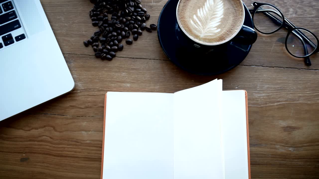 4K footage cinemagraph. blank white book on wooden table, page flip from wind. book at work space with notebook , cup of coffee and glasses