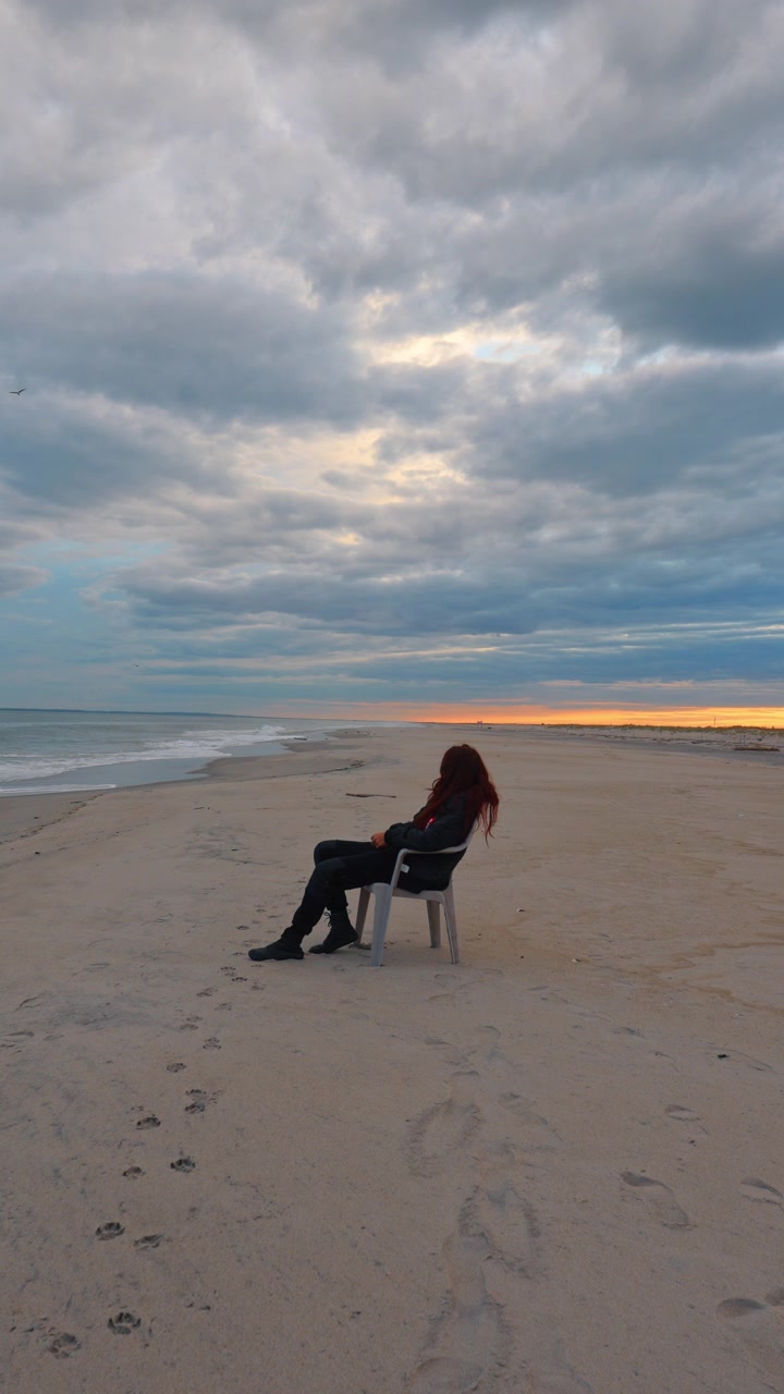 Person walking on a sandy beach near a plastic chair at dusk. Overcast clouds fill the sky above the sea horizon while foot tracks cover the shore sand.
