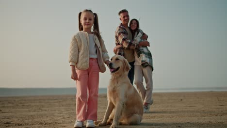 portrait of a happy little blonde girl in a white jacket and pink pants who stands near her big cream-colored dog and behind her her happy parents, a middle-aged man and a brunette girl in a green checkered shirt on a deserted seashore in summer