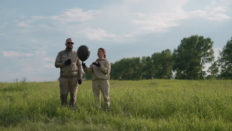 woman beside colleague in protective suits releasing black balloon while controlling it with handheld device, both standing in grassy field under blue sky with distant trees
