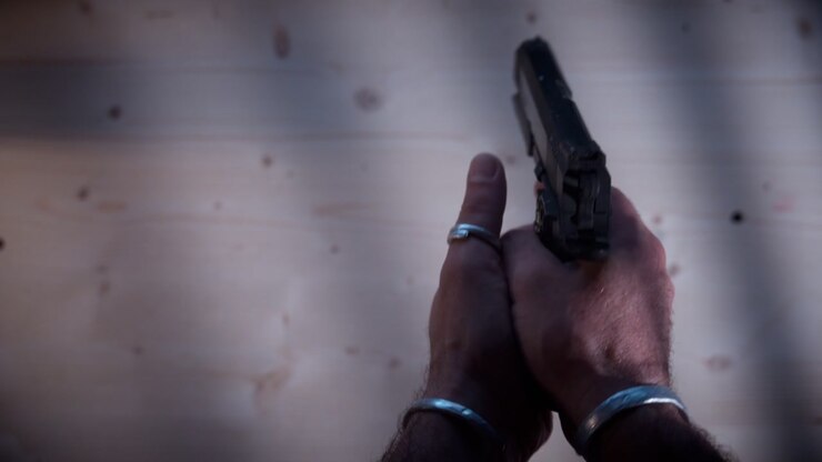 Primo piano di mani che tirano il carrello per camerare un proiettile in una pistola nera. Atmosfera da dark web sotterraneo con toni cinematografici cupi e prospettiva dall'alto.