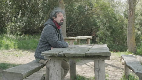 a middle aged man arriving at a picnic table in a forest, sitting down, taking a selfie