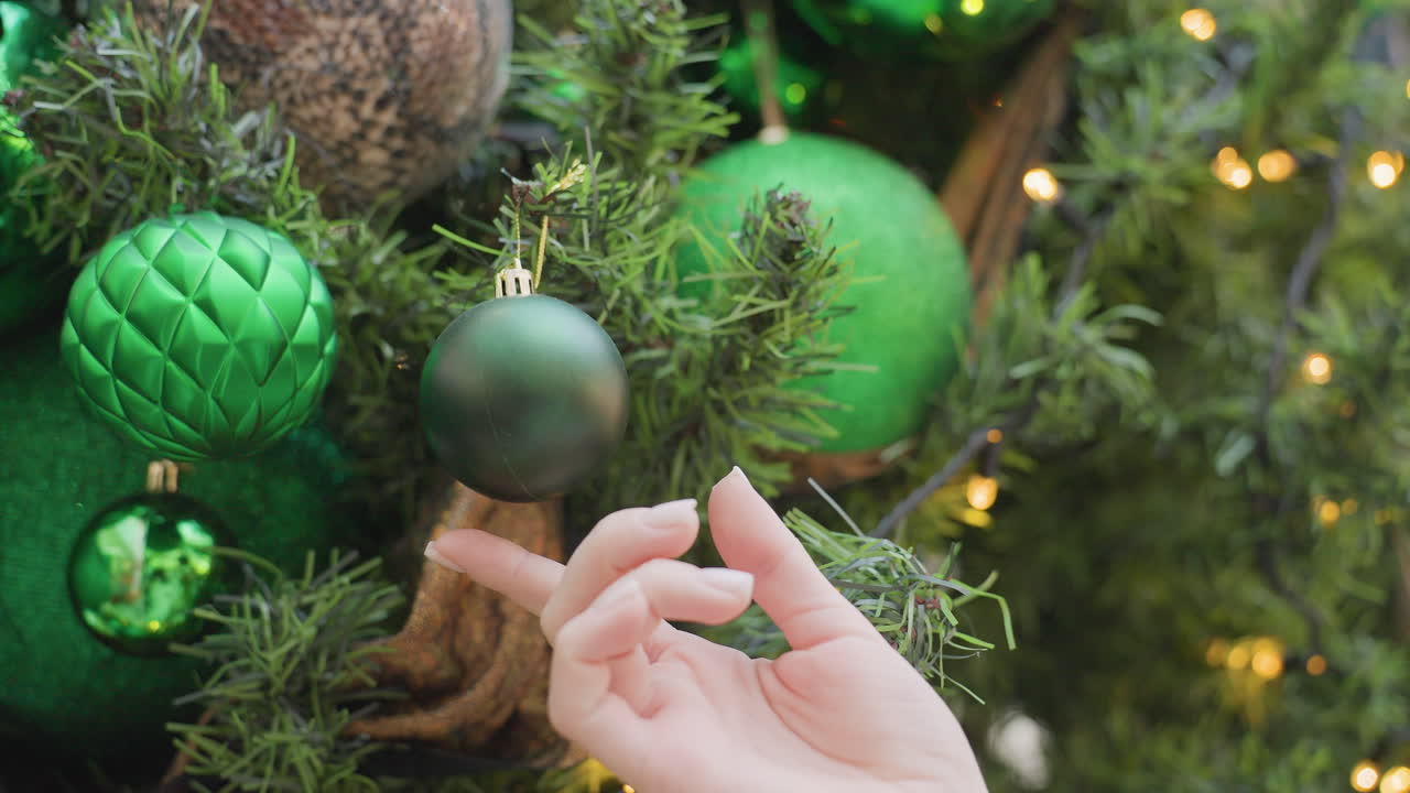 Close-up of hand holding small green decorative Christmas ball from tree, with lush greenery and soft, warm decorative lights shining