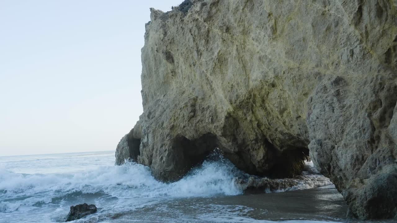 Powerful ocean waves crash against the base of a sea cliff, surging into a natural cave formation along a rugged coastline