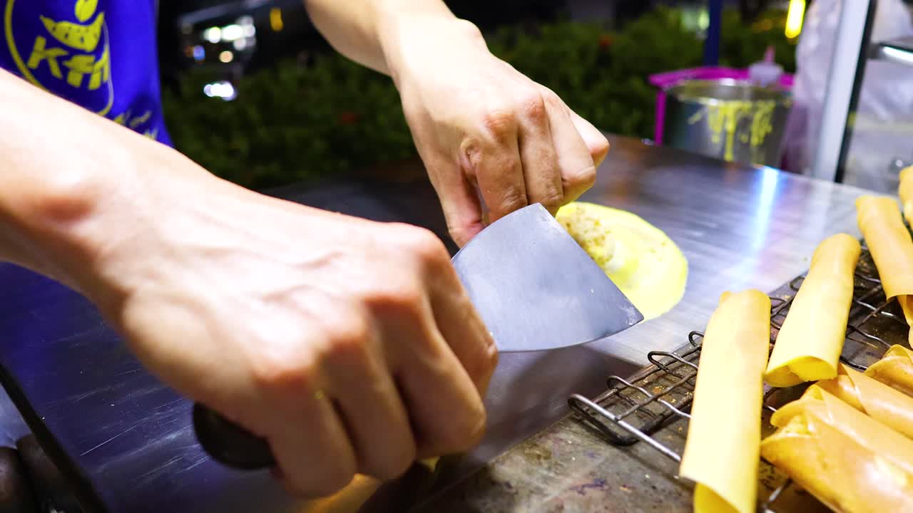 A street vendor skillfully prepares crispy pancakes on a metal surface under bright lighting in Bangkok, Thailand