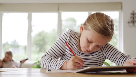 Niña-En-La-Mesa-Con-Tableta-Digital-Educación-En-El-Hogar-Durante-Una-Pandemia-De-Salud-Con-La-Familia-En-Segundo-Plano