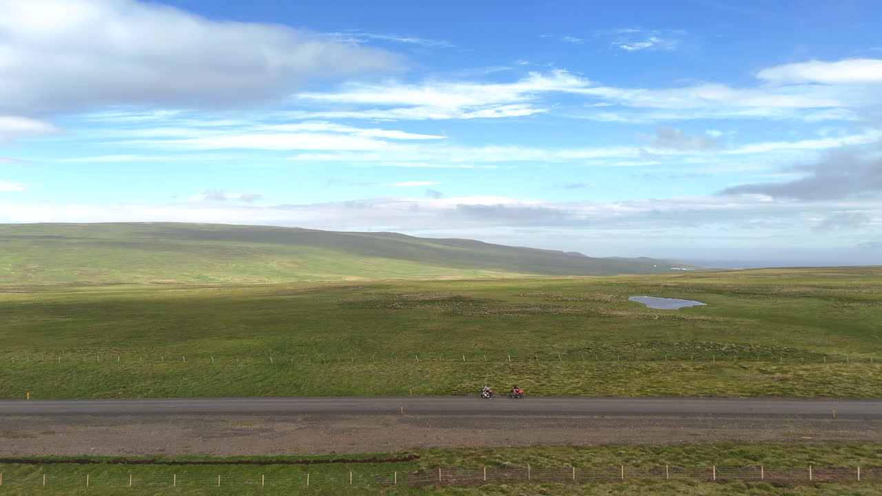 Luftfoto af to cyklister, der cykler på en afsidesliggende islandsk vej gennem åbne, grønne marker under en bred, blå himmel, der repræsenterer frihed, eventyrrejser og en bæredygtig udendørs livsstil.