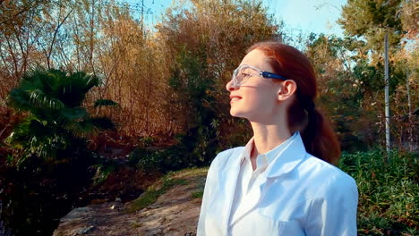 una joven científica en un arroyo, con gafas protectoras y una bata de laboratorio, mirando hacia la distancia y sonriendo con una mirada realizada