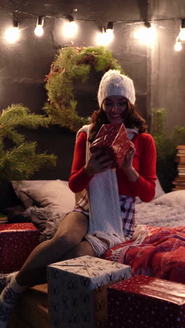 happy girl with gifts at christmas. attractive young woman in knitted white cap and scarf sitting on bed and looking at presents near new year tree at home. vertical video