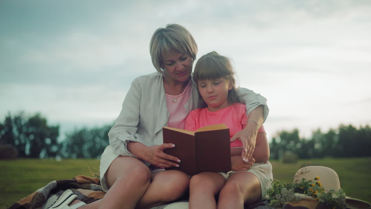 hermosa escena de una madre sosteniendo a su hija cerca, ambos sentados en el heno con un sombrero cercano, en un campo abierto al anochecer, compartiendo un libro