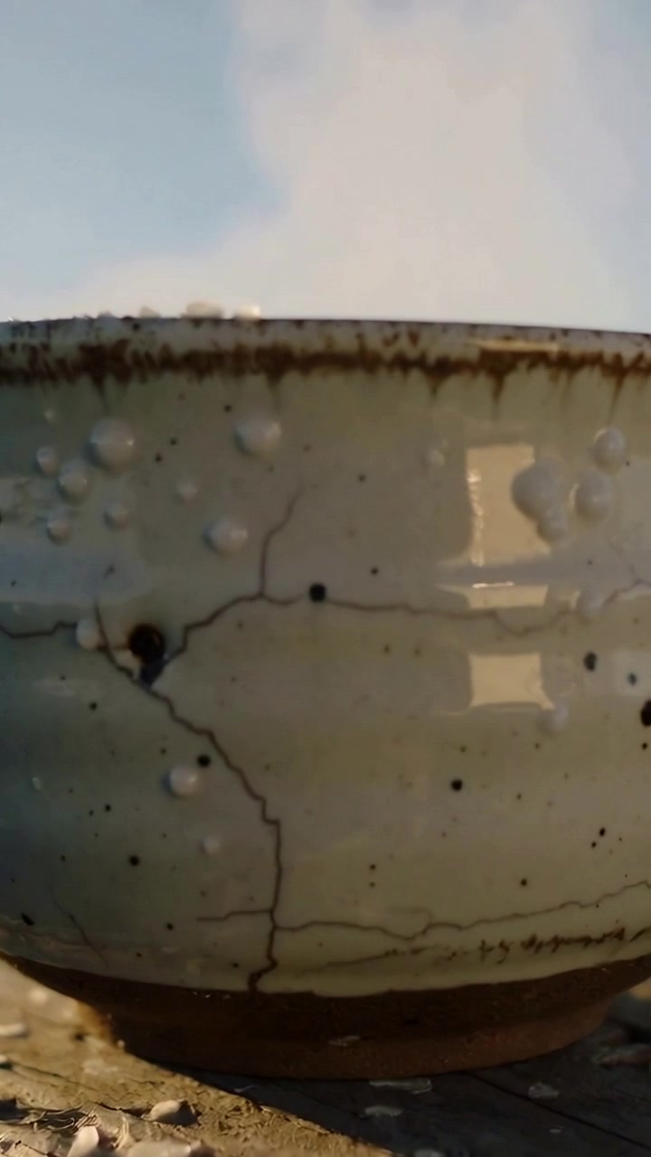 Vídeo vertical: Tigela de cerâmica esmaltada fumegante sobre tábua, luz do sol aquecendo o esmalte e levantando gotículas de água