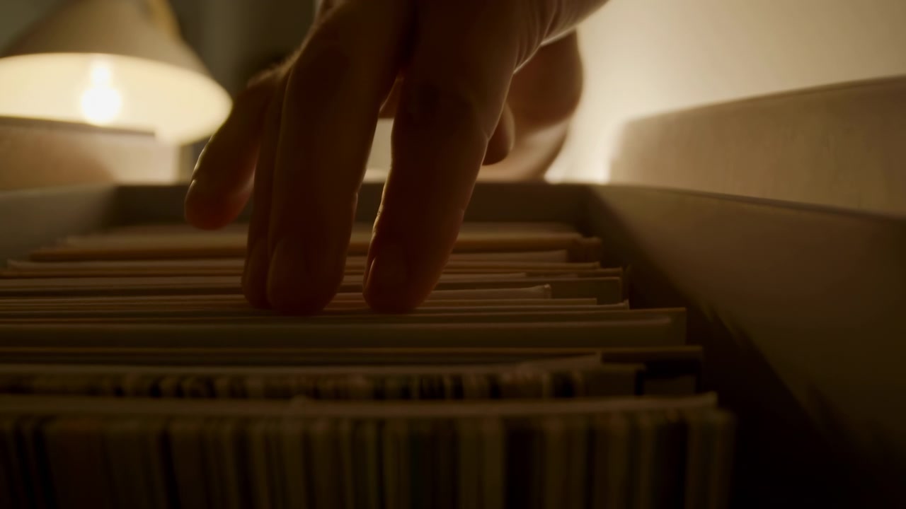 Una vista ravvicinata mostra una mano che si muove tra file di cartelle di file in un tranquillo archivio, con luce calda di una lampada, ombre morbide, messa a fuoco superficiale e documenti ordinati che suggeriscono un'attenta ricerca d'ufficio