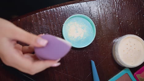 vista en primer plano de la mano de una joven que sostiene una esponja de maquillaje púrpura con polvo en ella, colocada dentro de un recipiente turquesa, demostrando la aplicación de maquilaje en la rutina de belleza