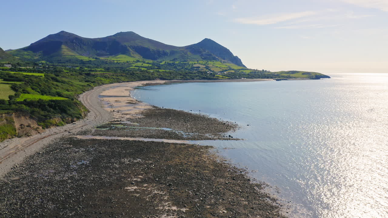 Drone flying over Trefor Beach and rugged Welsh coast in Wales with Yr Eifl Mountains in background on sunny day