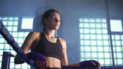 Young-tired-female-boxer-coming-to-the-corner-of-the-boxing-ring-and-resting-with-her-arms-on-the-ring-ropes-in-the-dark-gym