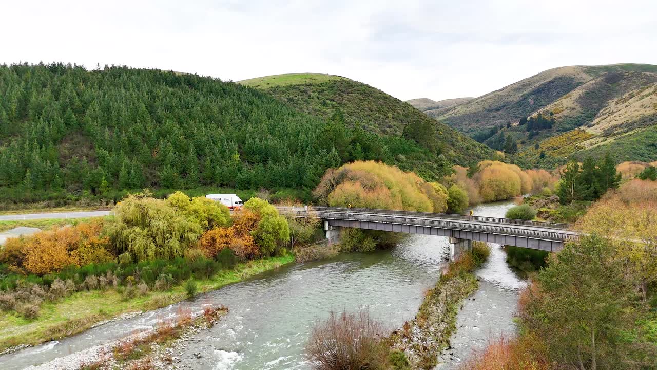 A vehicle crosses a bridge surrounded by lush greenery and hills under soft daylight at Lake Tekapo