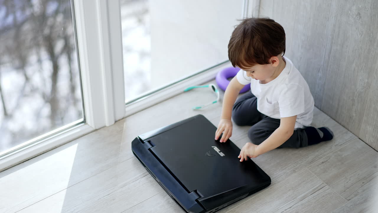 Caucasian toddler takes the laptop. Kid sitting on the floor opens the gadget. Top view.