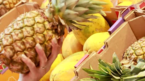 a person examines pineapples in a brightly lit supermarket, focusing on texture and ripeness among other tropical fruits