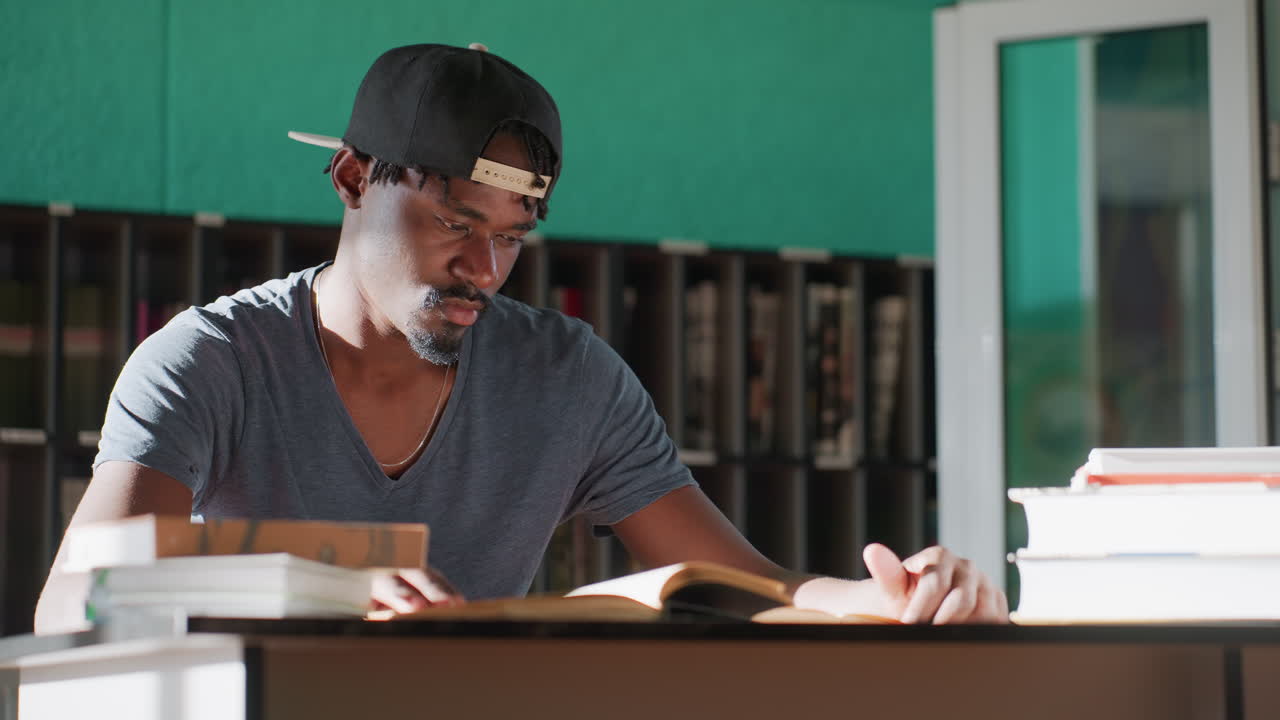 Remote worker in library studying, wearing cap, sitting at desk with stacks of books, tapping hand on table while reading, teal wall and shelves behind