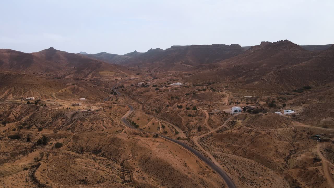 Aerial view of a winding road through the arid landscape of Ksar Hallouf, Tunisia. Sparse vegetation and scattered buildings dot the terrain.