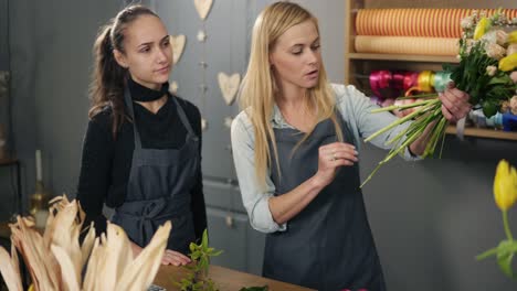 blonde florist in apron with her coworker at counter in floral shot while arranging counting the price for bunch of flowers