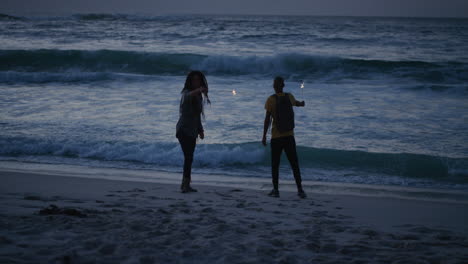 Amigos-Alegres-En-La-Playa-Bailando-Celebrando-La-Víspera-De-Año-Nuevo-Agitando-Bengalas-Disfrutando-De-Una-Diversión-Tonta-Juntos-En-Un-Tranquilo-Fondo-Marino-Junto-Al-Mar-Al-Atardecer