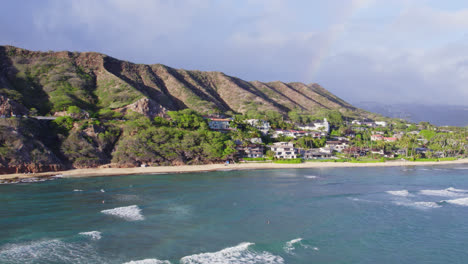 drone footage scanning the shore of the island of oahu and the volcanic mountain formations of diamond head and honolulu as a rainbow forms in the ocean mist