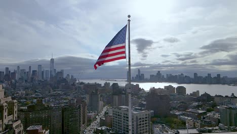 Toma-Aérea-En-Cámara-Lenta-De-La-Bandera-De-Estados-Unidos-Ondeando-En-El-Viento-Con-La-Ciudad-De-Nueva-York-En-El-Fondo