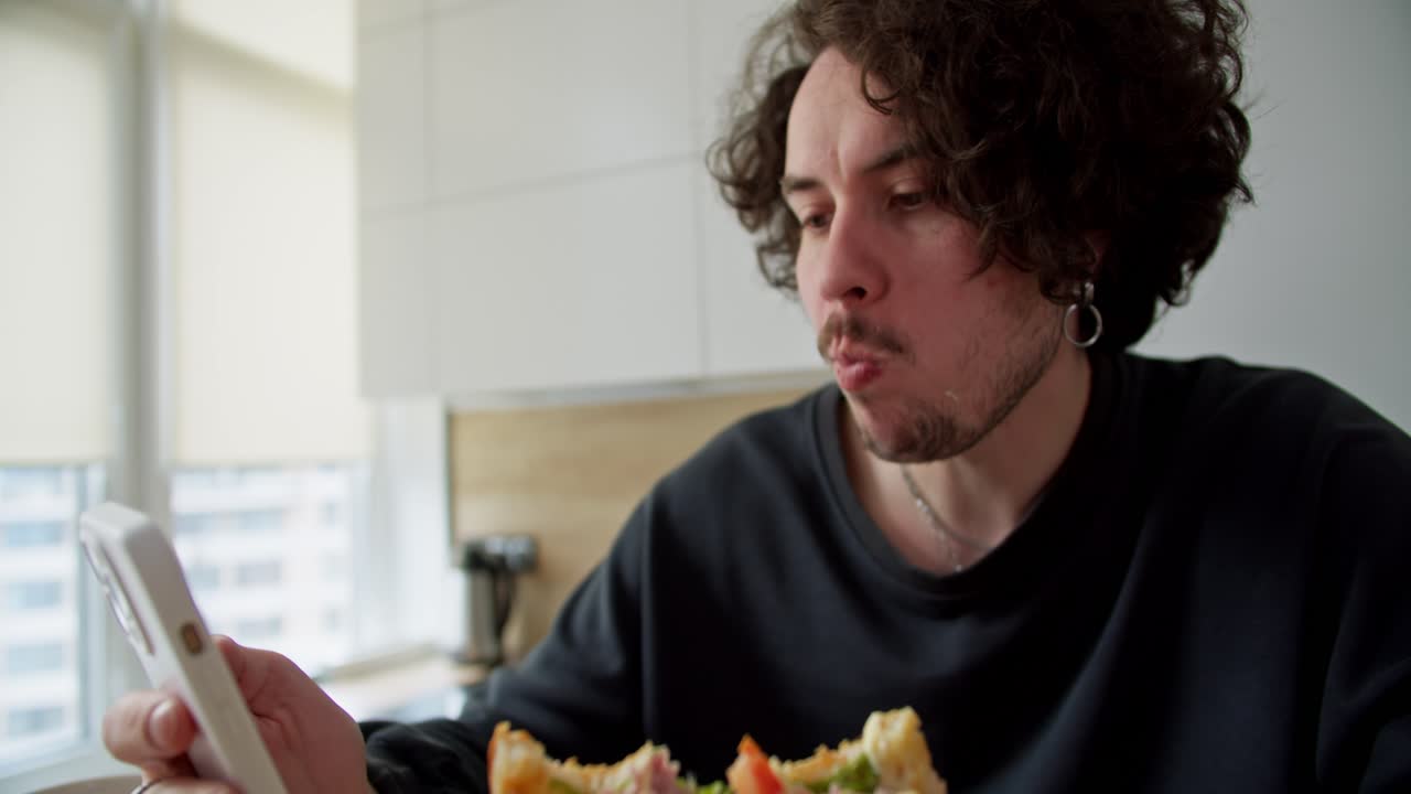 hombre moreno confiado con cabello rizado y bigote sentado y comiendo un sándwich mientras navega por las redes sociales en casa durante el desayuno en la cocina