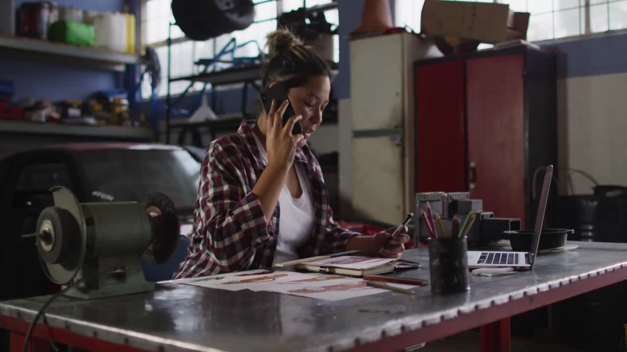 Meccanica donna che parla al telefono e usa un laptop in un'officina meccanica.