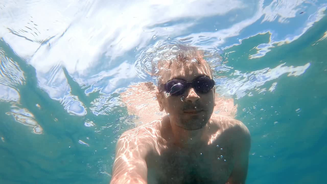 Man in swimming goggles under the water and holding camera, Aegean sea. Slow motion, underwater shooting. Greece