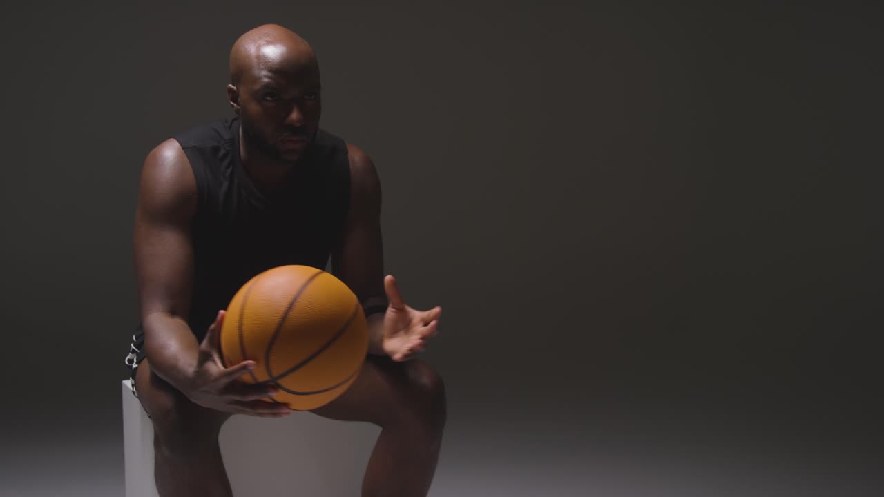 Studio Shot Of Seated Male Basketball Player With Hands Holding Ball