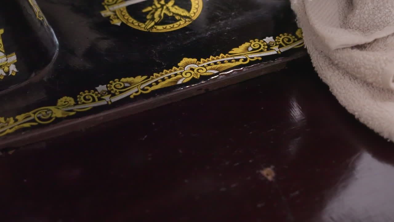 Una mano lucida la superficie di una macchina da cucire vintage, un panno leviga la decalcomania dorata ornata e la finitura laccata, delicata conservazione di un mobile d'epoca, un'attività di restauro concentrata in casa.