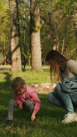 little girl and mother look for bugs in lawn grass in spring park. woman and daughter explore environment in urban garden. family walks in urban garden