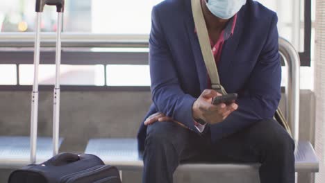 African-american-man-in-city-wearing-face,-waiting-for-bus,using-smartphone