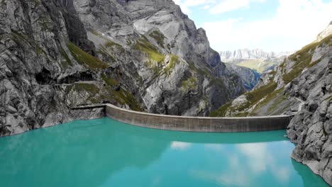 aerial flyover over turquoise waters of lake limmernsee and hydroelectric dam in linthal glarus, switzerland with cliffs and mountain peaks in background