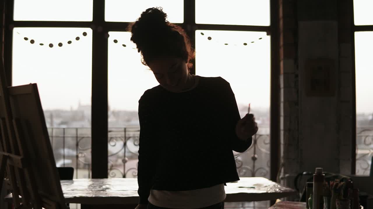 Young sweet female artist dancing while drawing her picture with paint brushes in her hands, in the loft styled art studio.