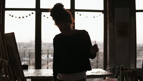 Young-sweet-female-artist-dancing-while-drawing-her-picture-with-paint-brushes-in-her-hands,-in-the-loft-styled-art-studio.