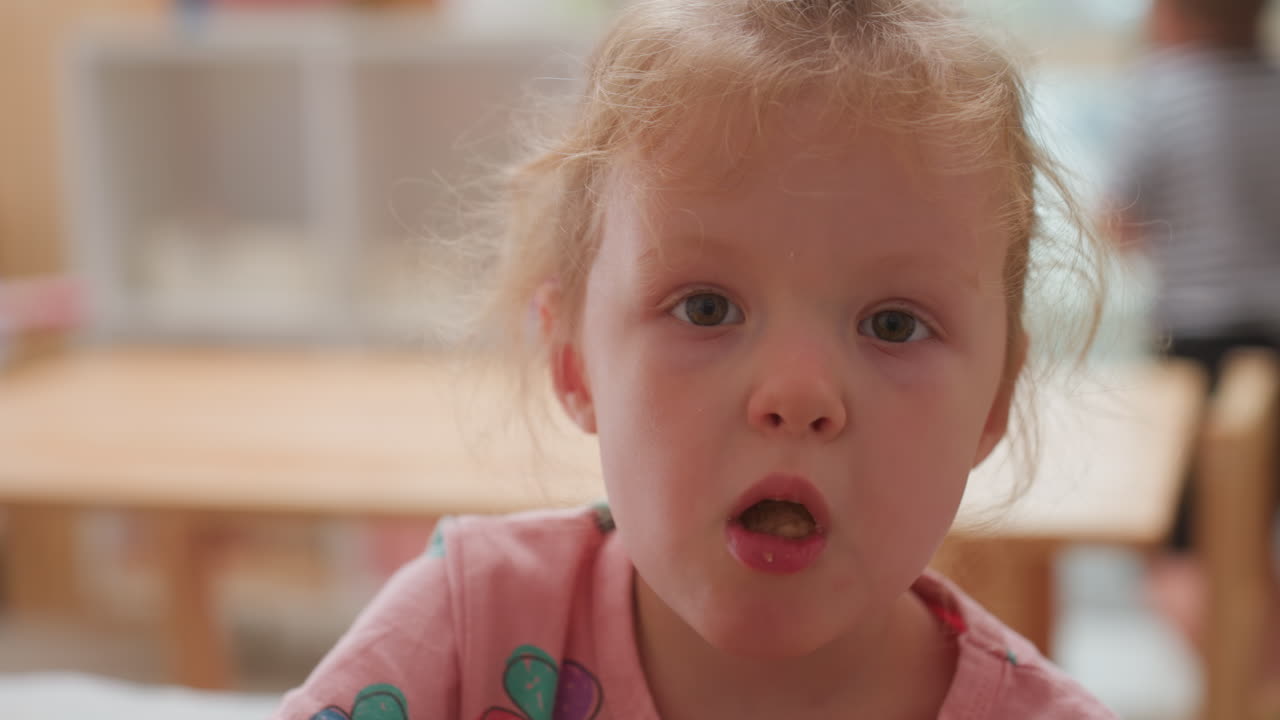 Close up of young girl with crumbs on lips looking toward camera after meal, soft daylight highlighting innocent expression, subtle curiosity and stillness
