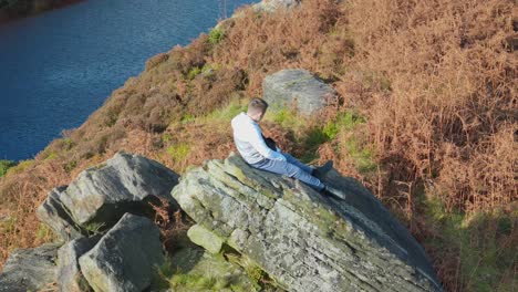 aerial video gently orbits a pensive young boy perched on a massive rock outcrop in the moorland setting